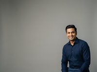 Young man with dark hair and olive complexion wearing navy blue button-up shirt, smiling at camera against neutral gray background. Suitable for corporate portraits, business profiles, professional headshots, and website content.