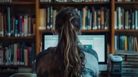 Young Asian woman with long dark hair working at computer in library setting, view from behind against wooden bookshelves filled with academic literature.