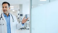 Professional male doctor with stethoscope presenting in modern hospital corridor, healthcare consultation and medical expertise concept.
