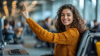 Young woman with curly dark hair wearing mustard sweater waves hello while working at modern office desk with laptop, expressing positive friendly attitude.