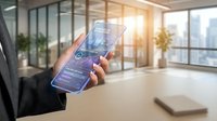 Businessman holding transparent smartphone with futuristic holographic dashboard interface in modern office, showcasing digital innovation.