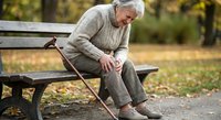 Senior Caucasian man with walking cane sitting on park bench in autumn, experiencing loneliness and contemplation during golden hour outdoor setting.
