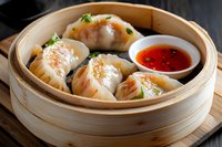 Steamed dumplings with shrimp filling in bamboo basket served with spicy chili sauce, garnished with fresh herbs on wooden table, traditional Asian cuisine.