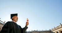 Mature male graduate in academic regalia and mortarboard gesturing while celebrating outdoors near classical university architecture on clear day.