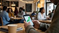 Person with dark skin tone holding smartphone displaying green checkmark in casual café setting with diverse patrons and warm lighting, suitable for technology, success, approval, and business communication concepts.