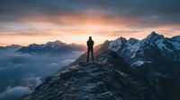 Silhouette of hiker standing on mountain peak at sunrise with dramatic clouds and snow-capped alpine ranges in background creating inspirational landscape scene.