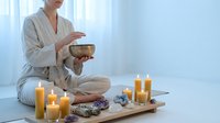 Woman in beige robe meditating with singing bowl surrounded by candles and crystals in serene spa setting for wellness and mindfulness practices.