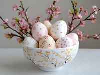 Decorated Easter eggs with delicate pink patterns in elegant white ceramic bowl with gold floral design, alongside blooming cherry blossom branches on light surface.