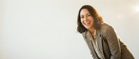 Young woman with dark hair and olive complexion wearing a casual beige blazer, smiling warmly while looking at camera against neutral background. Suitable for business, lifestyle, and professional website use.