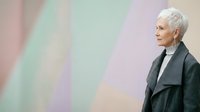 Senior woman with short white hair, Caucasian ethnicity, wearing dark gray blazer over white top, standing in profile against pastel striped background. Suitable for lifestyle, aging, fashion, and corporate concepts.