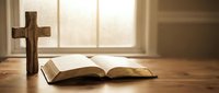 Wooden cross and open bible on table with warm sunlight streaming through window creating peaceful spiritual atmosphere for prayer and meditation.