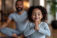 Young child with curly hair and medium skin tone practicing yoga or meditation with hands in prayer position, smiling with eyes closed. Adult male in background in casual home setting, promoting wellness and mindfulness for families.