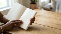Person with dark skin tone holding open blank notebook on wooden table near window, suitable for education, journaling, writing, planning, and business concepts.