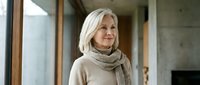 Mature woman with long white hair and fair complexion wearing a beige turtleneck sweater, standing indoors by a window in a modern home setting.