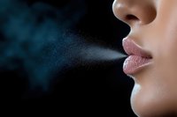 Close up of feminine lips exhaling fine mist or vapor against dark background, showing delicate droplets suspended in air creating atmospheric effect.