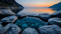 Tranquil mountain lake at sunset with smooth rocks framing crystal clear water revealing submerged stones, dramatic landscape with soft orange sky reflection.