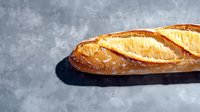 Fresh crusty French baguette with golden brown crust on gray concrete background with harsh shadow, traditional artisanal bread photography.