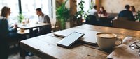 Modern workspace with smartphone and coffee cup on wooden table in busy cafe with blurred people working in background for business productivity concepts.