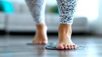 Woman stepping on bathroom scale barefoot wearing patterned leggings for weight loss fitness and health monitoring at home.