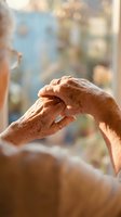 Elderly hands with age spots gently touching in warm sunlight by window, showing connection, care and tenderness between senior couple in golden hour lighting.