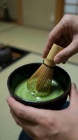 Caucasian male hands whisking matcha green tea powder in black ceramic bowl with traditional bamboo whisk during Japanese tea ceremony preparation.