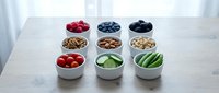 Fresh healthy snacks arranged in white bowls on marble counter featuring berries nuts vegetables and fruits for clean eating lifestyle.