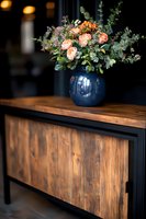 Fresh peach garden roses and eucalyptus arrangement in navy blue ceramic vase on rustic wooden console table with dark metal frame, moody interior setting.