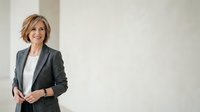 Middle-aged woman with short blonde hair wearing dark blazer and white shirt, standing against neutral background for corporate and business use.