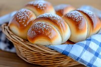 Fresh homemade dinner rolls with sesame seeds in wicker basket lined with blue checkered napkin, golden brown crust and soft texture, close up view.