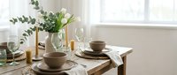 Elegant dining table setting with ceramic dinnerware, eucalyptus branches in vase, wine glasses and natural light from window creating cozy atmosphere.