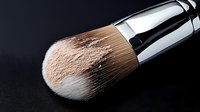 Professional makeup brush with loose powder particles on black background, close-up view showing soft bristles and chrome handle for cosmetic application and beauty tutorials.
