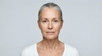 Confident mature Caucasian woman with gray hair wearing white top against neutral background for skincare and wellness campaigns.