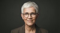 Senior woman with short white hair and eyeglasses smiling at camera against dark background. Professional portrait for business, healthcare, education, and corporate communications.