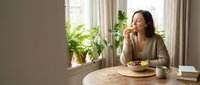 Woman with long dark hair and medium skin tone sitting at wooden table eating healthy salad near window with plants, suitable for wellness, nutrition and lifestyle content.