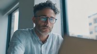 Middle-aged man with glasses and curly gray hair looking concerned while working on laptop in modern office space with city view through window.