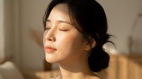 Young East Asian woman with eyes closed in serene expression, experiencing relaxation, wellness, or skincare routine in warm indoor lighting.