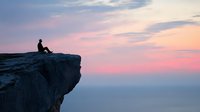 Silhouette of person sitting alone on cliff edge against dramatic pink and purple sunset sky, contemplating vast horizon over misty mountain landscape.