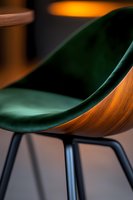 Modern curved chair with emerald green velvet upholstery and wooden shell on black metal legs, close-up view showing luxurious texture and warm lighting.