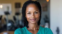 Young African American businesswoman with braids and bright smile wearing turquoise blouse in office setting, radiating confidence and professionalism.