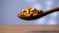 Mixed nuts including cashews, almonds, and hazelnuts on wooden spoon for healthy snacking and cooking ingredients on neutral background.