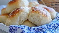 Fresh homemade dinner rolls with golden brown tops dusted with flour, served in woven basket with blue and white patterned napkin, close up view of soft bread texture.