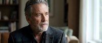 Senior man with gray hair and beard wearing dark gray cardigan, looking thoughtfully to the side in home interior setting for lifestyle and portrait use.