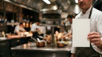 Chef in white chef's coat holding blank white card in professional kitchen with staff working in background, suitable for restaurant menus, culinary promotions, and food service advertising.