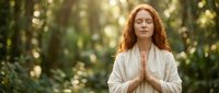 Young redhead woman in prayer pose meditating peacefully in sunlit forest setting with soft natural lighting and bokeh background for wellness content.