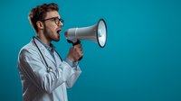 Young male doctor with glasses shouting into megaphone on blue background for healthcare communication and medical announcement campaigns.
