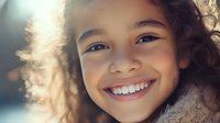 Young mixed race girl with curly hair and bright genuine smile showing white teeth, glowing in soft natural light against blurred background.