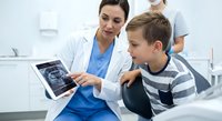 Female dentist showing dental X-ray results to young Caucasian boy patient on tablet in modern clinic office for oral health consultation and treatment.