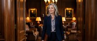 Professional blonde businesswoman in navy suit standing confidently in elegant wood-paneled office with warm lighting and classic furniture.