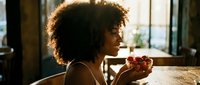 Young woman with curly auburn hair and medium skin tone enjoying fresh strawberries at wooden table indoors, casual lifestyle moment for food and wellness content.