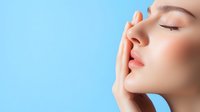 Young Asian woman with natural makeup touching face against blue background, eyes closed, showing clean glowing skin for beauty and skincare advertising.